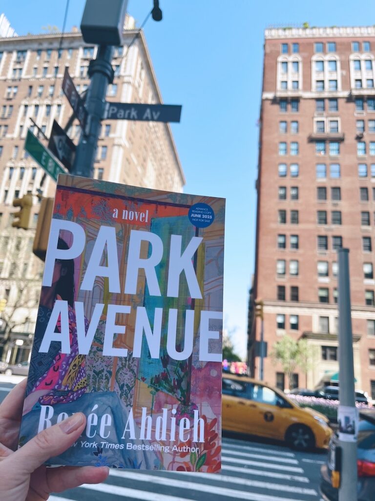 A hand holding a copy of Park Avenue at the corner of Park Avenue. Buildings line the avenue and a yellow taxi whizzes by